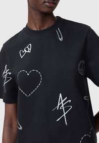 Mustat puuvillat-paita, jossa on valkoisia graafisia kuvioita, kuten sydämiä, turvaneuloja ja rauhan symboleja. Lyhyet hihat ja pyöreä kaula-aukko.