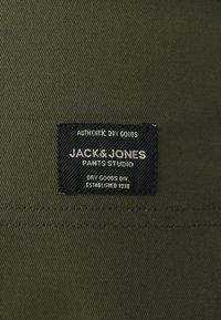 Maslina zelena tkanina s teksturiranom površinom koja sadrži crnu oznaku s natpisom "JACK & JONES PANTS STUDIO" u bijelom slovu.