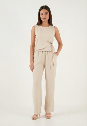 Nő áll, bézs színű ujjatlan felsőt visel oldalt megkötve, hozzá illő laza nadrágot és bézs színű magassarkú szandált egy sima fehér háttér előtt.