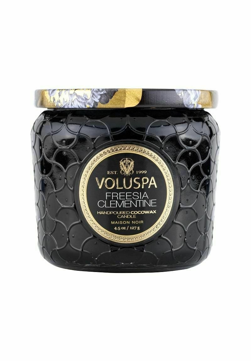 Svart, texturerad ljusbehållare med ett lock som har gulddetaljer. Etiketten visar "Voluspa Freesia Clementine", 4,5 oz, handgjord cocowax.