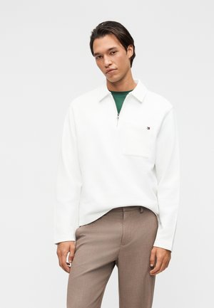 Jeune homme portant un pull blanc zippé par-dessus une chemise verte et un pantalon beige, debout devant un fond clair et uni.