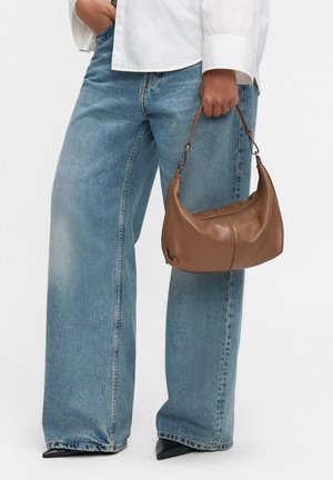 Borsa a tracolla in pelle marrone con una forma curvata e cuciture sottili, tenuta da una persona. Abbinata a jeans larghi di colore blu chiaro e scarpe nere a punta.