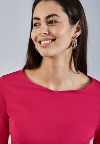 Jasnoróżowa bluzka z szerokim dekoltem i długimi rękawami, w połączeniu ze srebrnymi kolczykami w kształcie kół. Gładki materiał z delikatnym połyskiem.