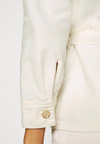 Manga de chaqueta blanca cremosa, con cierre de botón y ligero plisado. Textura suave con sutil patrón de tejido diagonal.