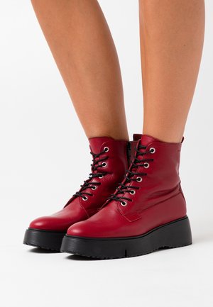 Bottes courtes en cuir rouge à lacets avec semelles épaisses en caoutchouc noir, portées sur des jambes nues sur fond blanc.