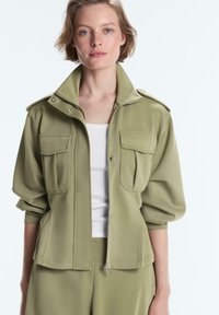 Frau trägt eine olivgrüne Jacke mit großen Klappentaschen über einem weißen gerippten Tanktop, stehend vor einem schlichten hellen Hintergrund.