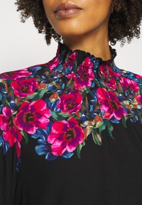 Un primo piano di un colletto con un motivo floreale, con fiori rosa e blu vivaci su uno sfondo nero. Il modello ha i capelli ricci.