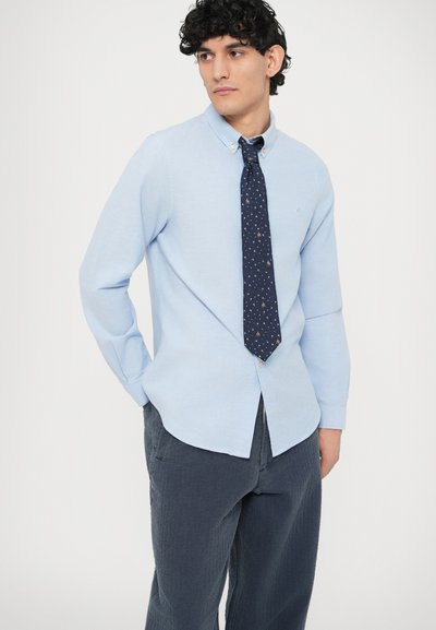 Ανοιχτό γαλάζιο πουκάμισο με κουμπιά και γιακά, συνδυασμένο με σκούρη γραβάτα με σχέδια. Το μοντέλο φορά σκούρο παντελόνι με υφή και ποζάρει με τα χέρια πίσω από την πλάτη.
