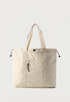 Beige tote bag with two long fabric handles, front zip pocket with pull cord, drawstring sides, and snap closure at the top.