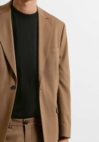 Hombre vestido con una chaqueta de traje color beige y pantalones a juego sobre una camiseta negra de cuello redondo, cortada en el cuello y la cintura, contra un fondo blanco.