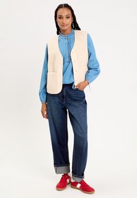 Gilet en polaire beige avec deux poches avant sur une chemise en denim bleu clair, associé à un jean foncé retroussé et des baskets rouges avec des accents blancs.