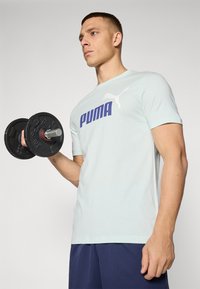 Šviesiai mėlyna trumpomis rankovėmis marškinėliai su dideliu purpuru "PUMA" logotipu. Asmuo laiko juodą hantelį su apvaliais svarmenimis, vilkėdamas tamsiai mėlynas šortus.