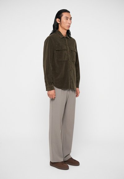 Camisa de veludo em verde-azeitona, com fecho de botões à frente e dois bolsos no peito, combinada com calças largas cinza-claro e sapatos castanhos.