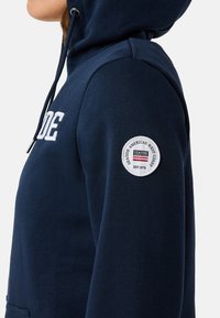Sweatshirt marine avec capuche, arborant un logo brodé blanc sur le devant et un écusson circulaire sur la manche. Tissu texturé.