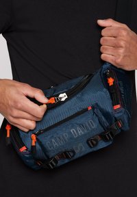 Blaues Stoff-Slingbag mit orangefarbenen Akzenten, mehreren Reißverschlusstaschen, schwarzem Zubehör und "CAMP DAVID" auf der Vorderseite gedruckt. Strukturiert Oberfläche.