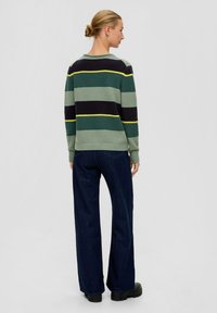 Frau mit blonden Haaren im Dutt, die einen grün, schwarz und gelb gestreiften Pullover, dunkelblaue Jeans und schwarze Stiefel trägt, steht vor einem schlichten Hintergrund.