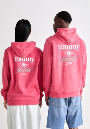 Felpe con cappuccio rosa dal taglio rilassato, con testo del logo bianco e blu chiaro e una grafica sul retro. L'abbigliamento da uomo è abbinato a pantaloni corti in denim.
