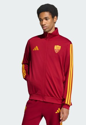 Joven con chaqueta y pantalones deportivos rojos y amarillos con logos del equipo y de Adidas, de pie con las manos en los bolsillos.