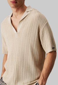 Camisa polo de manga corta color beige con un diseño de canalé texturizado, que presenta un escote en V y un cuello en contraste. Logotipo sutil en la manga.