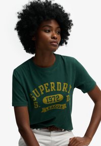 Zielony bawełniany t-shirt z żółtym napisem „SUPERDRY” oraz „1972 LEAGUE.” Okrągły dekolt, krótkie rękawy, luźny krój.