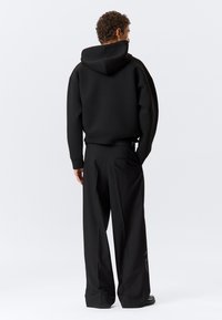 Μαύρο φούτερ με κουκούλα και άνετη εφαρμογή, σε συνδυασμό με μαύρο παντελόνι φαρδιάς γραμμής. Το ύφασμα φαίνεται μαλακό και λείο με ορατές λεπτομέρειες υφής.