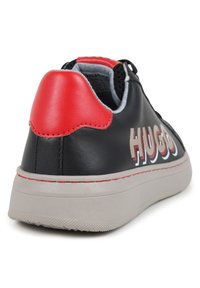Sneaker en cuir noir avec un accent de talon rouge, semelle grise, et lettrage "HUGO" bien en vue sur le côté. Présente une surface texturée.