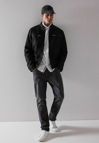 Czarna kurtka z zamkami na kieszeniach noszona na białej koszuli, w połączeniu z ciemnymi dżinsami i białymi trampkami. Model swobodnie opiera się o neutralne tło.