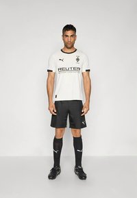 Λευκή φανέλα ποδοσφαίρου με μαύρες λεπτομέρειες, με λογότυπο και κείμενο χορηγού. Συνδυάζεται με μαύρο σορτς, μαύρες κάλτσες και παπούτσια με τάπες.