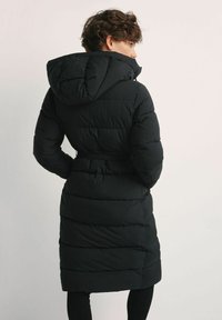 Długi czarny pufferski płaszcz z kapturem, z paskiem w talii i poziomym pikowaniem. Posiada gładką, błyszczącą teksturę.