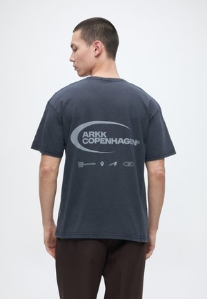 Άνδρας με την πλάτη, φοράει σκούρο γκρι μπλούζα ARKK Copenhagen με λογότυπο και ιστοσελίδα τυπωμένα στο πίσω μέρος, μπροστά σε απλό φόντο.