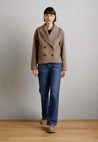 Veste en laine marron croisée avec de grands revers et deux boutons, associée à un jean en denim bleu et des chaussures grises. Texture lisse, coupe décontractée.