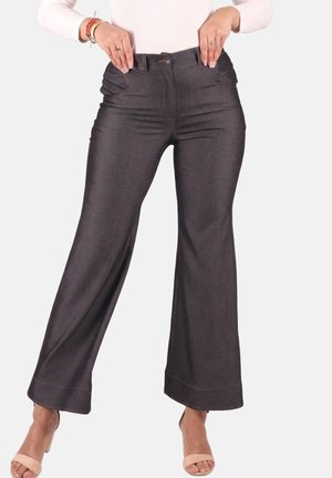 Frau trägt dunkel ausgestellte Hose und beige offene Absätze, die Hände liegen nahe am Bund, vor einem schlichten hellen Hintergrund.