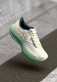 Hellbeige und grüne Lauf Schuhe in der Luft über einer Asphaltoberfläche, die die strukturierte Sohle und die Logo-Details an der Seite zeigen.