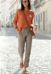 Rost-orange gestrickter Pullover, hellbeige Jogginghose mit Kordelzug, braune flache Sandalen und eine beige Umhängetasche. Helles Sonnenlicht, Kopfsteinpflasterstraße.