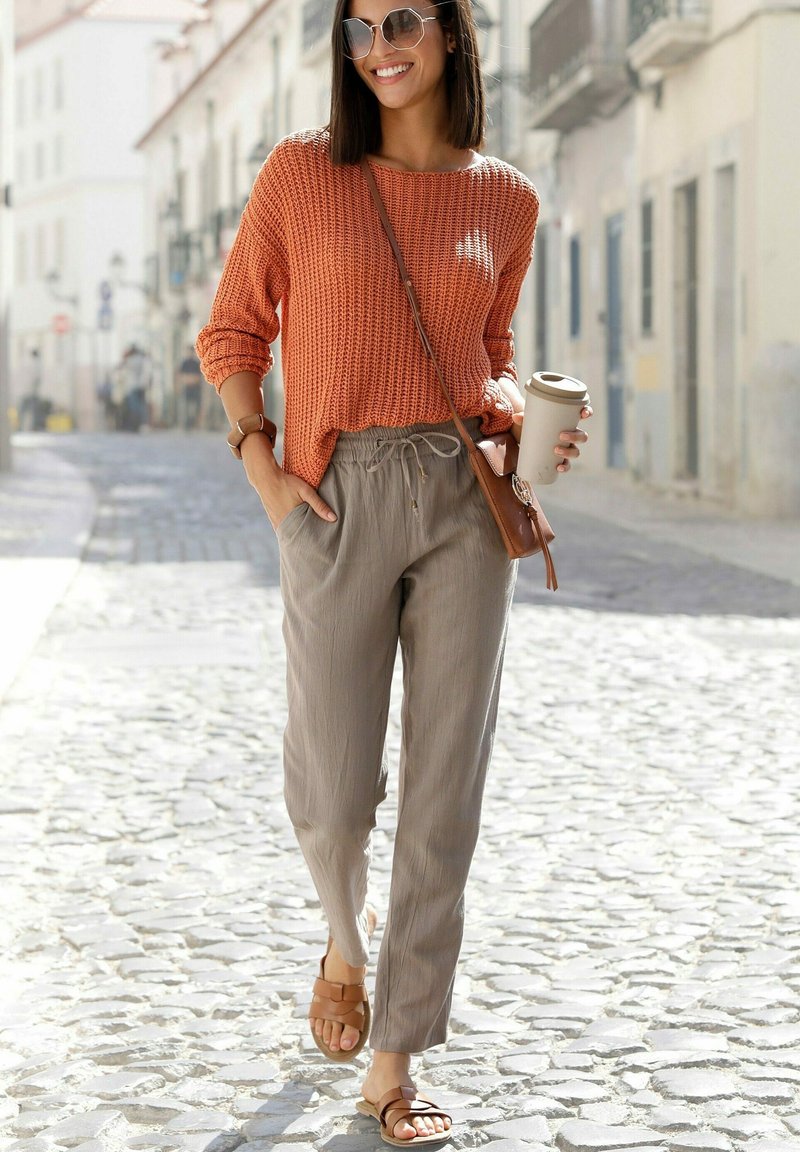 Rost-orange gestrickter Pullover, hellbeige Jogginghose mit Kordelzug, braune flache Sandalen und eine beige Umhängetasche. Helles Sonnenlicht, Kopfsteinpflasterstraße.