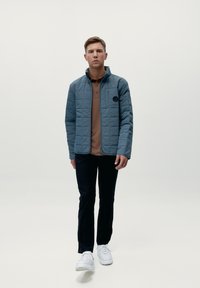 Chaqueta azul acolchada con cremallera frontal y cuello alto, llevada sobre una camiseta polo marrón, combinada con pantalones negros y zapatillas blancas.
