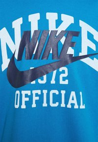 Turkos Nike-t-shirt med stora vita och marinblå grafiker, inklusive varumärkesnamnet, året 1972 och ordet "Official."