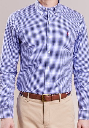 Mann trägt ein blau-weiß kariertes Hemd mit Knöpfen, das in beigen Hosen mit einem braunen Ledergürtel steckt.