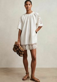 Vrouw staat in een witte korte jurk met kanten uitsneden op de mouwen en de zoom, terwijl ze een geweven tas vasthoudt en tan enkelband sandalen draagt.