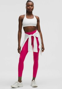 Witte sportbh, strakke roze leggings en een witte blouse om de taille geknoopt. Model draagt witte sportschoenen, effen achtergrond.