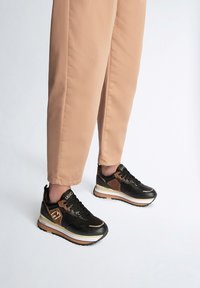 Scarpe sportive nere e marroni con finitura lucida, dotate di una suola bianca spessa, lacci e dettagli di marca. Indossate con pantaloni beige a tinta unita.