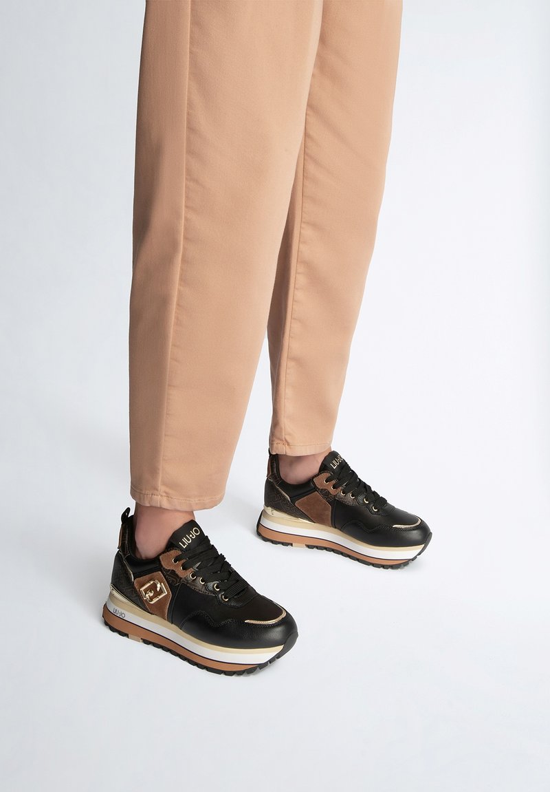 Scarpe sportive nere e marroni con finitura lucida, dotate di una suola bianca spessa, lacci e dettagli di marca. Indossate con pantaloni beige a tinta unita.