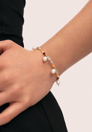 Perlenarmband mit goldenen Akzenten, das weiße Perlen, mehrfarbige Edelsteine und ein zartes Design aufweist. Am Handgelenk getragen.