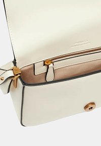 Bolso de cuero crema con herrajes dorados, un bolsillo con cremallera y un forro marrón en contraste. Presenta un diseño estructurado y detalles en negro.