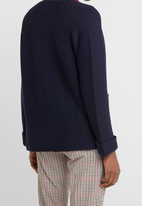 Cardigan blu navy con texture e vestibilità ampia, con polsini arrotolati e design rilassato. Indossato sopra dei pantaloni a quadri.