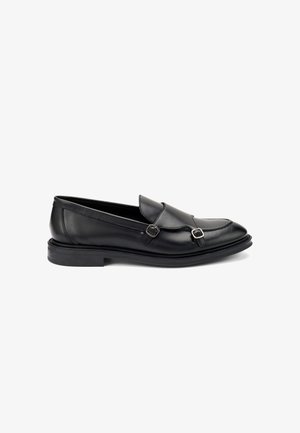 Los mocasines de cuero negro presentan una punta redondeada, acentos de doble hebilla y una suela de goma con un ligero tacón. Textura suave y diseño clásico.