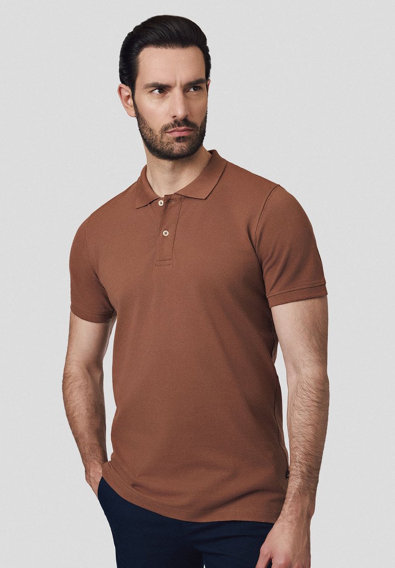 Brązowa koszulka polo z krótkim rękawem, klasycznym kołnierzykiem i trzema guzikami, uszyta z gładkiego materiału, noszona z granatowymi spodniami.