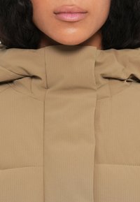 Giacca imbottita in tessuto beige con una sottile texture a coste. Presenta un colletto alto e dettagli di cucitura nascosti per un design elegante.
