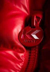 Röd puffjacka av tyg med en slät textur. Har en plastdragkedja och ett rundat draghandtag märkt med en stiliserad "K" och "JK11T."