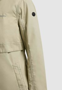 Manche de veste beige avec bouton rond noir sur le haut du bras et poche inclinée cousue en dessous, couture visible et logo partiel sur le côté gauche.